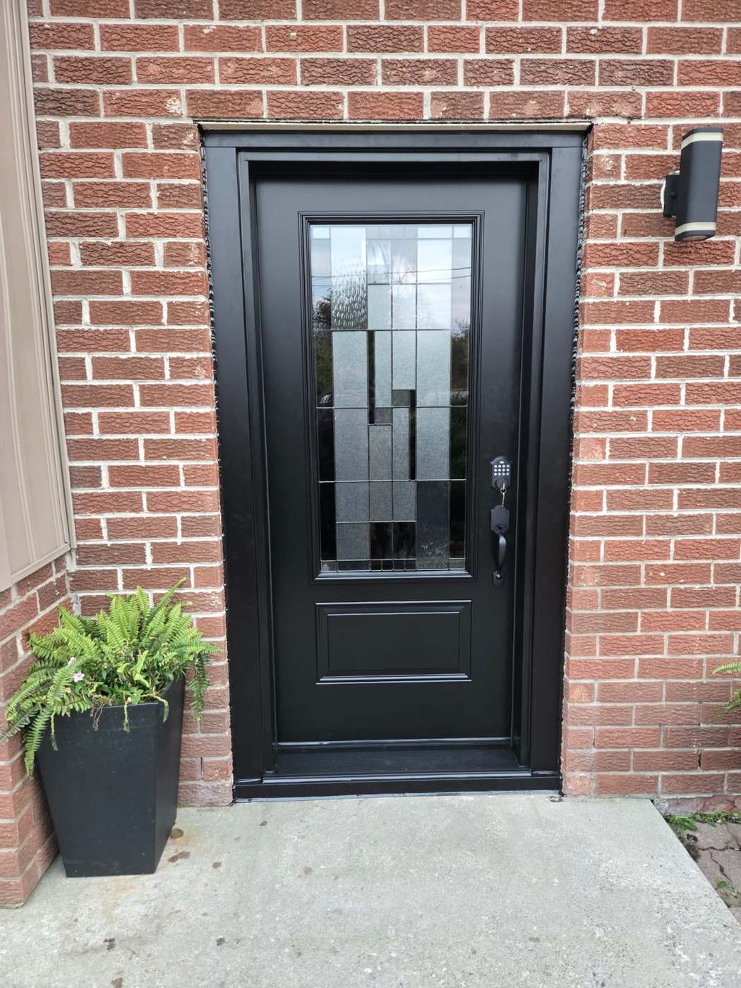 Black steel entrance door with decorative geometric glass insert and keypad deadbolt on a red brick home